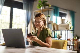 Frau sitzt zu Hause am Tisch mit offenem Laptop und Smartphone in der Hand